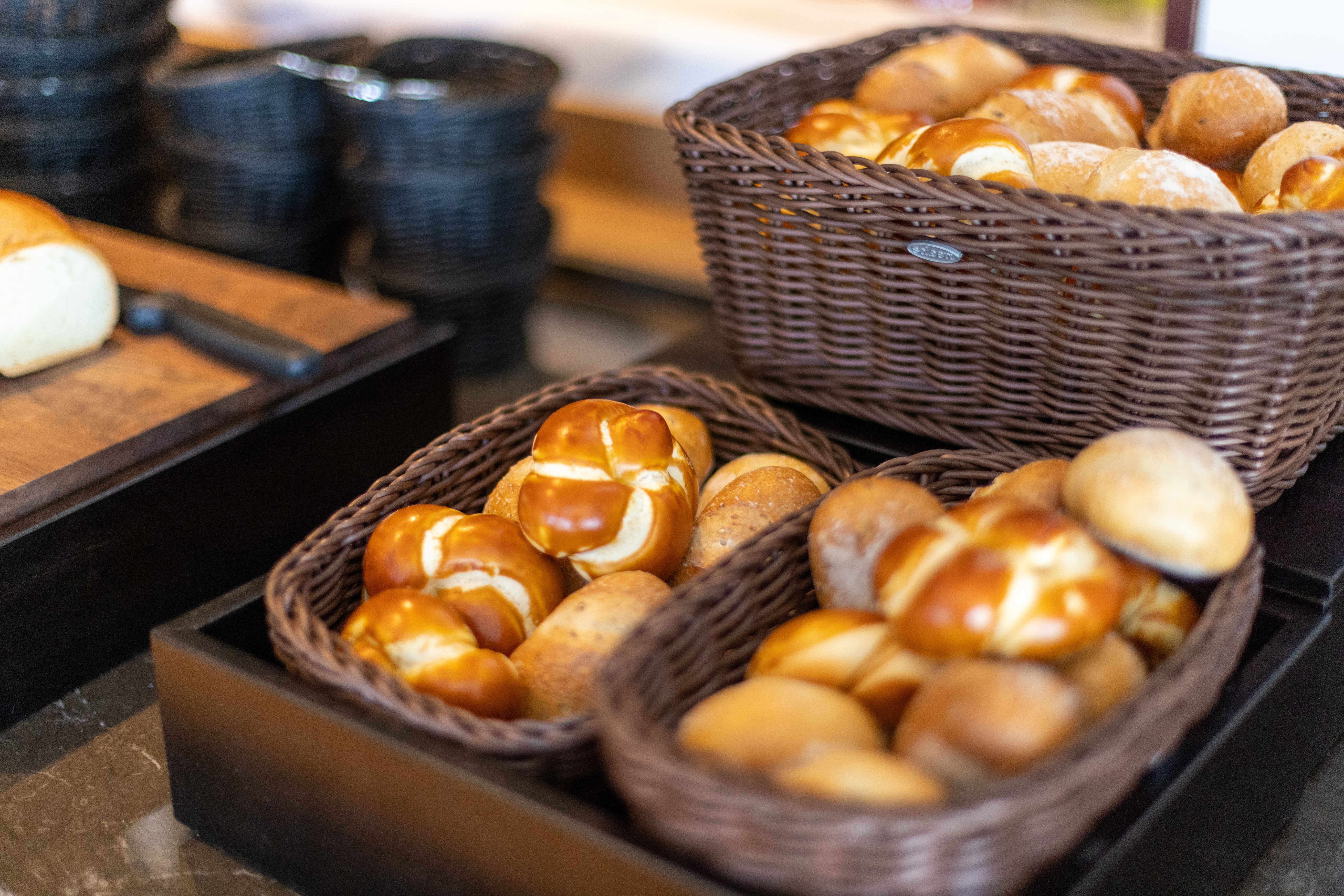 Mehrere gefüllte Körbe mit frischen Backwaren, darunter goldbraune Brötchen und hellere Brotsorten. Geflochtene braune Körbe, teils auf Tabletts. Im Hintergrund ein Gefäß und Brot auf einem Schneidebrett. Brunch-Buffet.